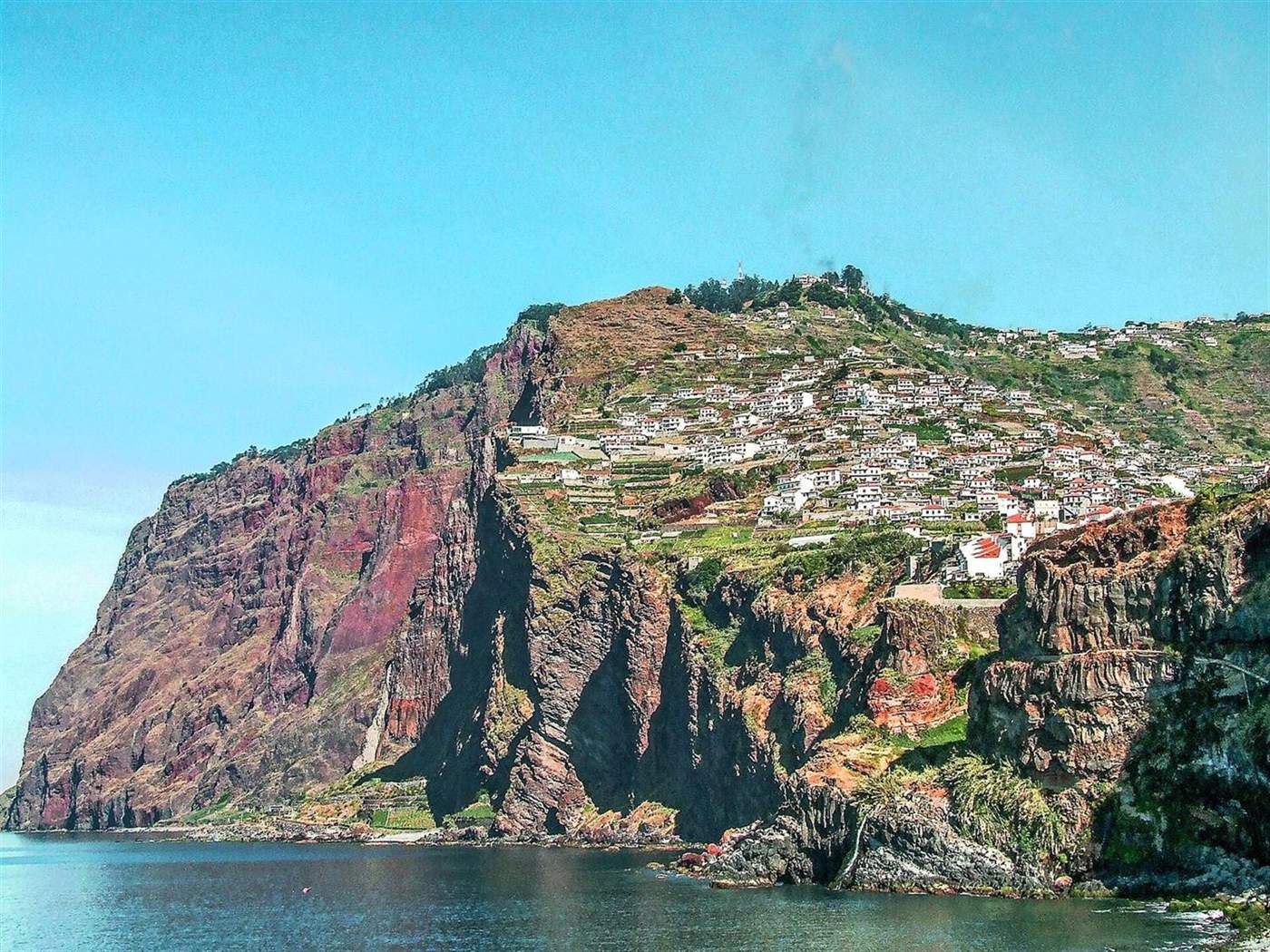 Steep red-brown cliffs of Cabo Girão with a hillside village on top, seen from the sea near Câmara de Lobos