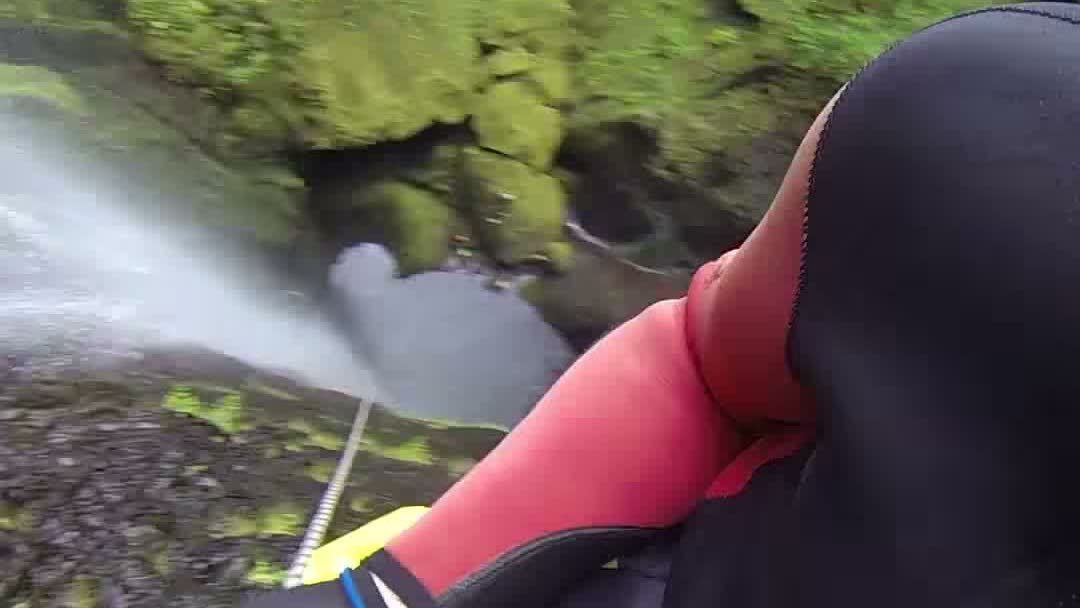 Canyoner in a wetsuit and harness rappelling past a waterfall on a rope at Ribeira da Hortelã, Madeira