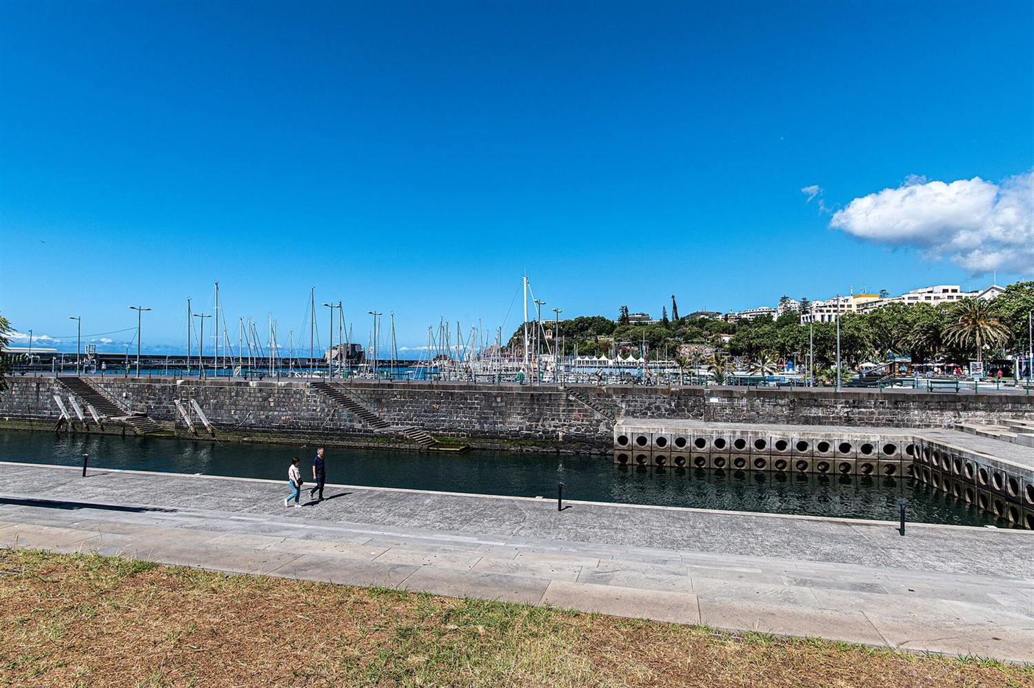 Marina and stone harbour walls with two people walking along the promenade in Funchal, Madeira