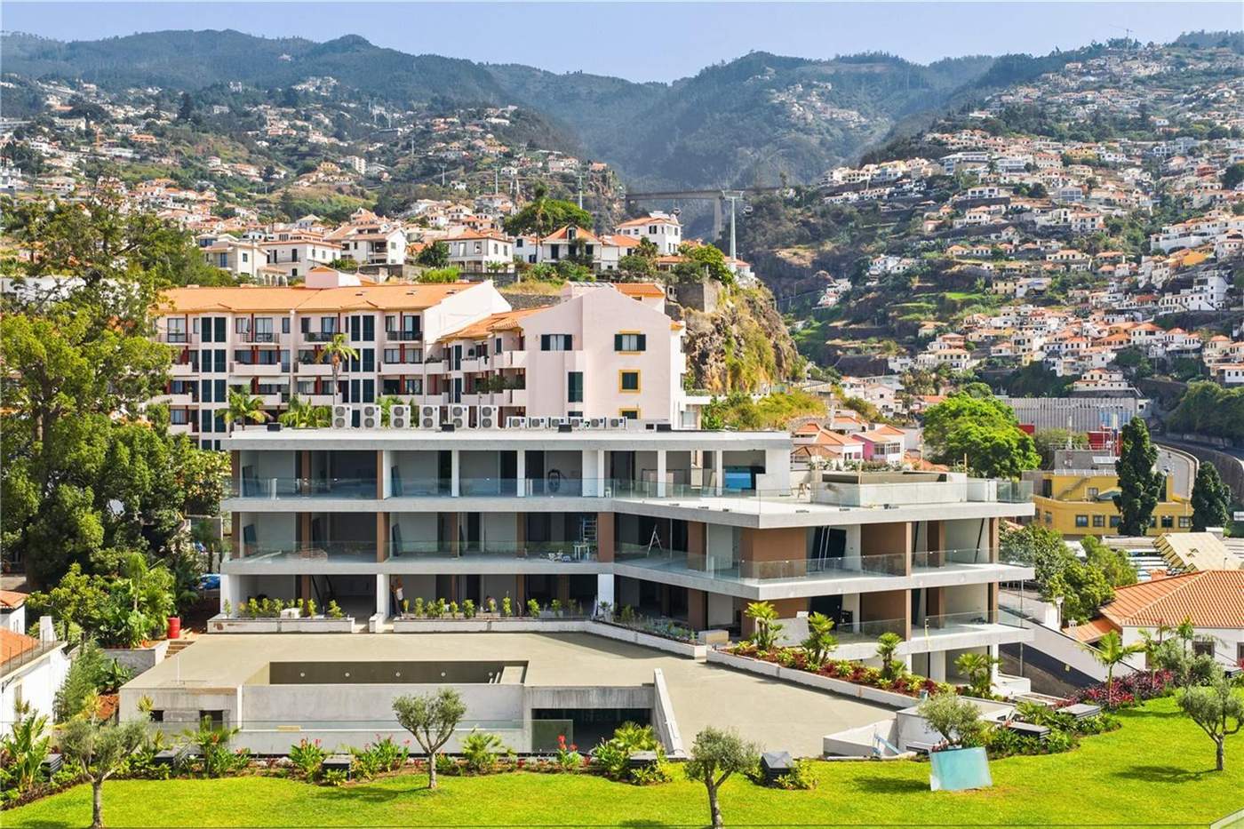 Aerial view of the Uptown Lux blocks, long white balconied buildings with lawns in front and terracotta-roofed Funchal houses climbing the green hillside behind