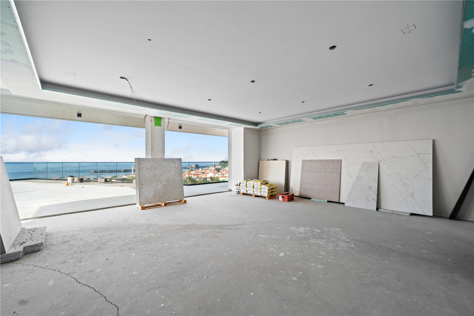 The main living space unfinished, floor-to-ceiling glazing along the sea side and stone slabs stacked against the wall