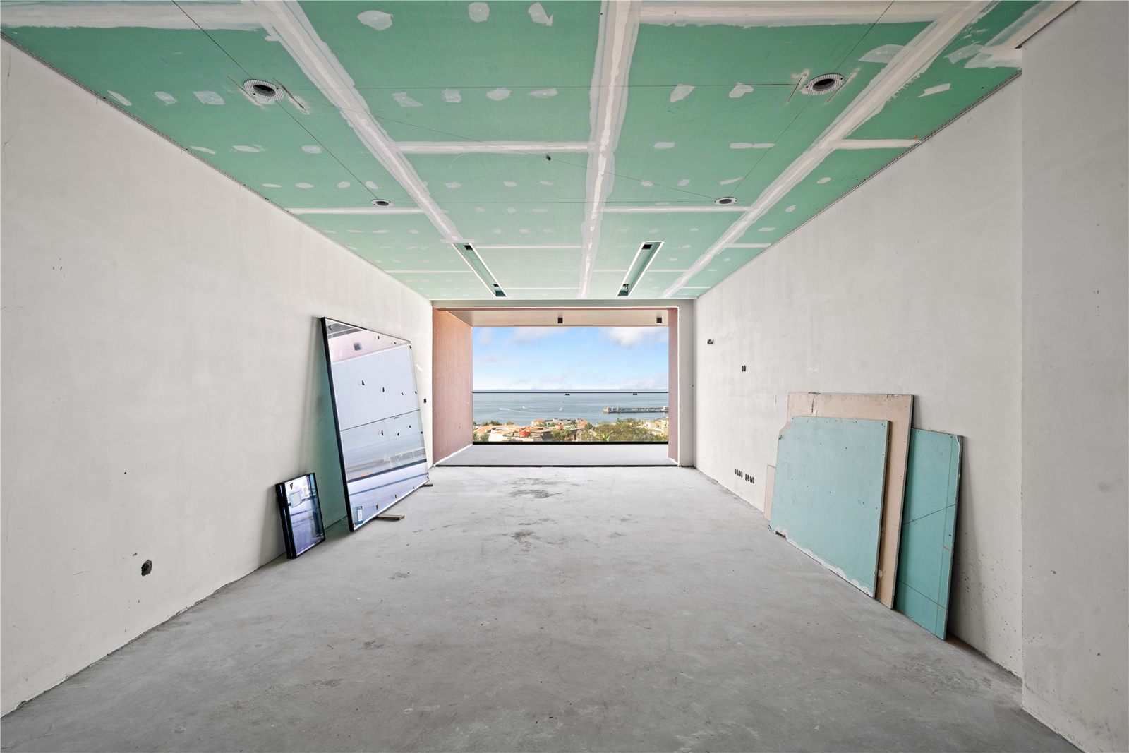 The living space under construction, bare concrete floor and a wide opening to the balcony with the sea beyond