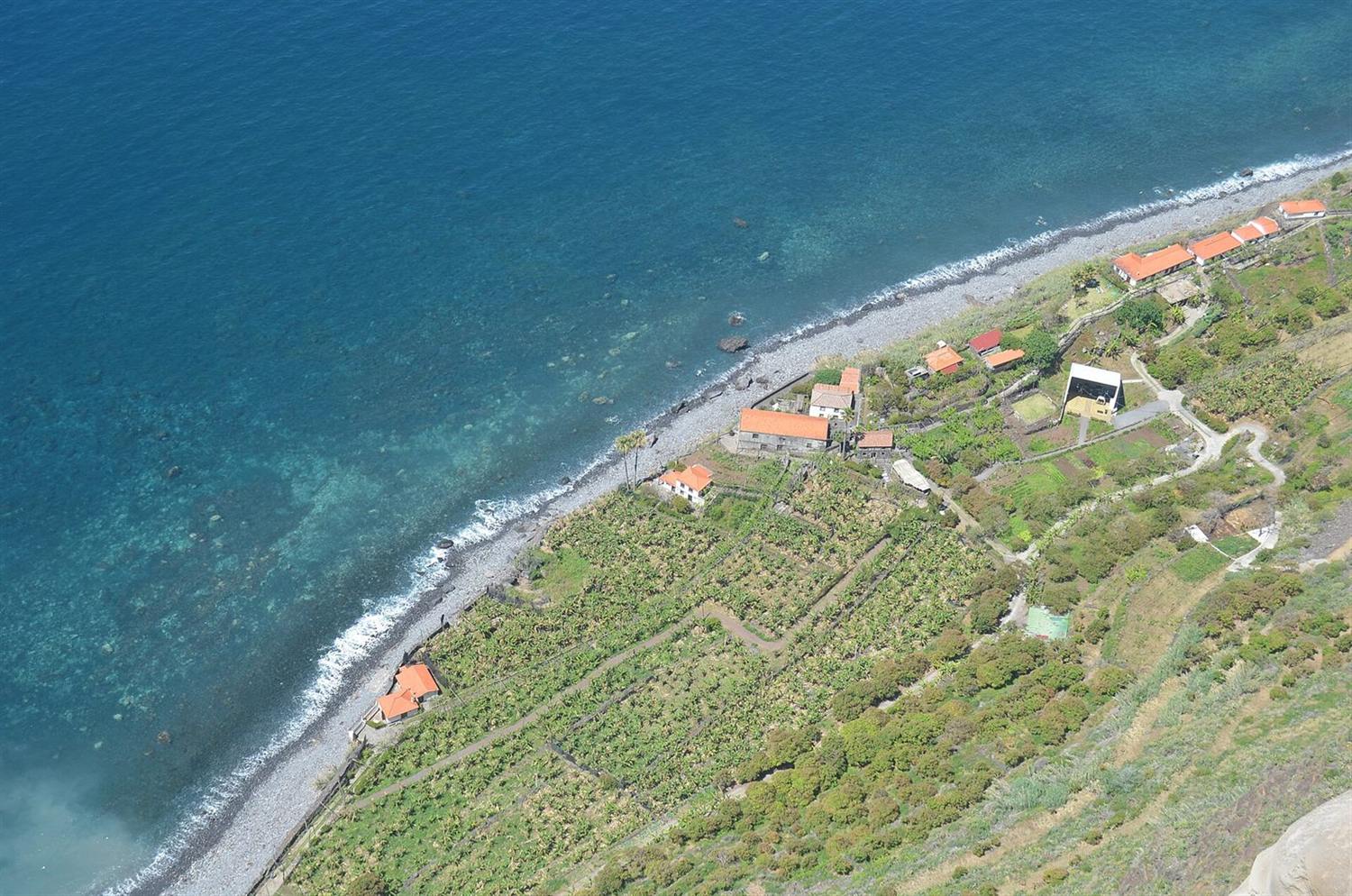 Aerial view of a coastal terraced farmland with orange-roofed houses along a pebble beach in Madeira