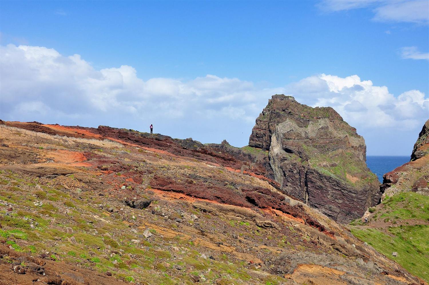 Red and ochre volcanic ridge at Ponta de São Lourenço, Madeira, with a lone hiker and sea beyond