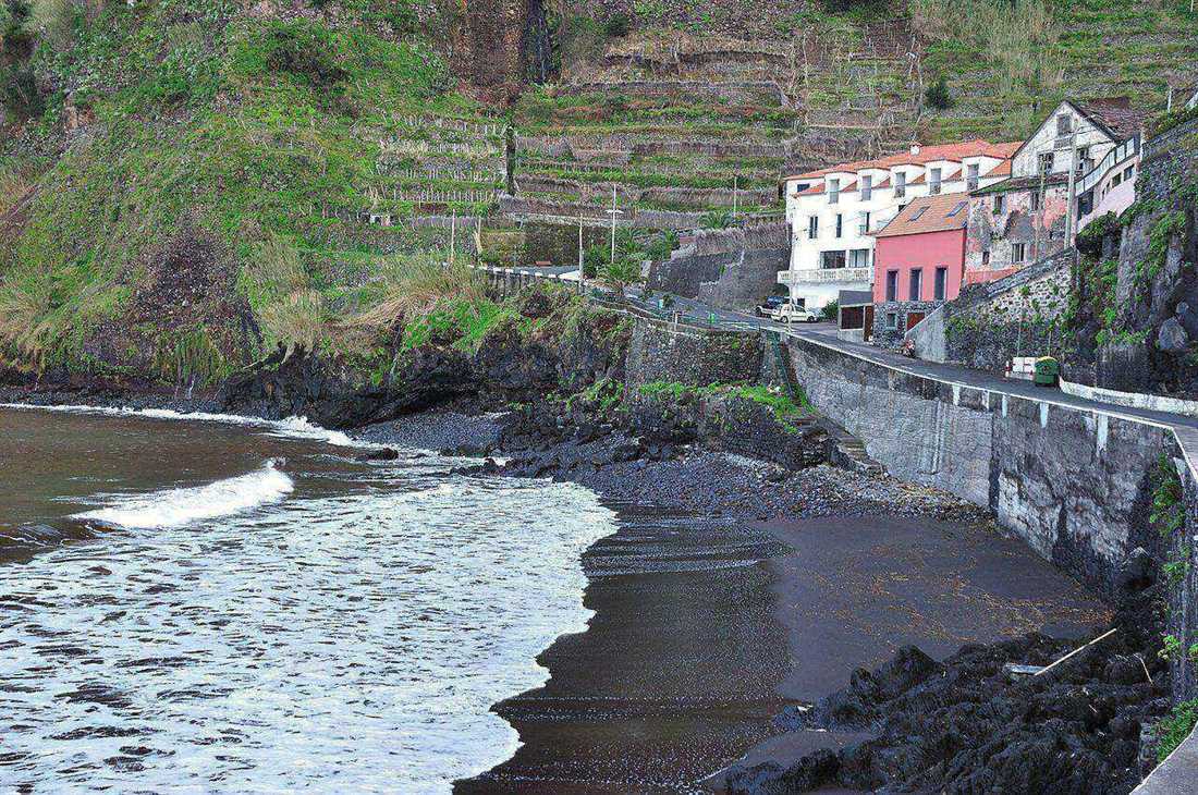 Black volcanic sand beach at Seixal, with terraced vineyards on the cliff behind and houses along the coast road, Madeira