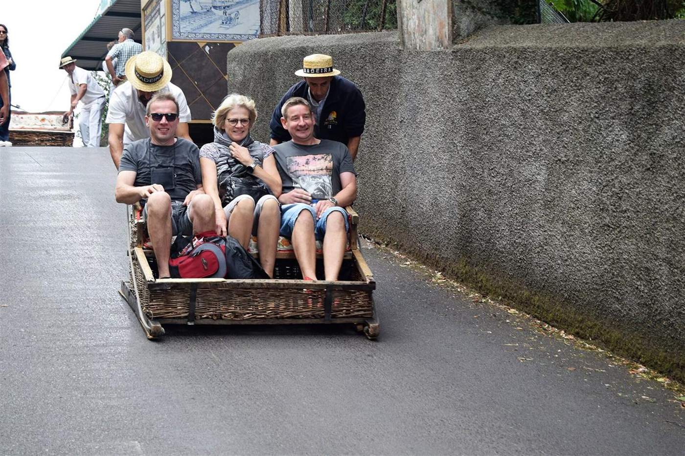 Wicker toboggan sledge carrying three smiling passengers, pushed by two hat-wearing drivers in Monte, Funchal, Madeira